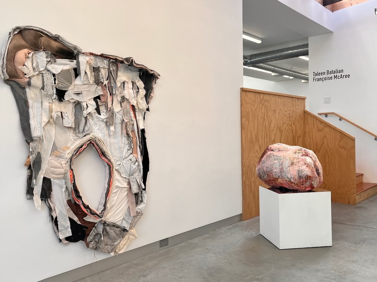 Taleen Batalian's multimedia work hangs on a white wall to the left, while McAree's pink bulbous sculpture sits on a pedestal to the right. In the back, there are stairs leading up to the show at Overlap Gallery.