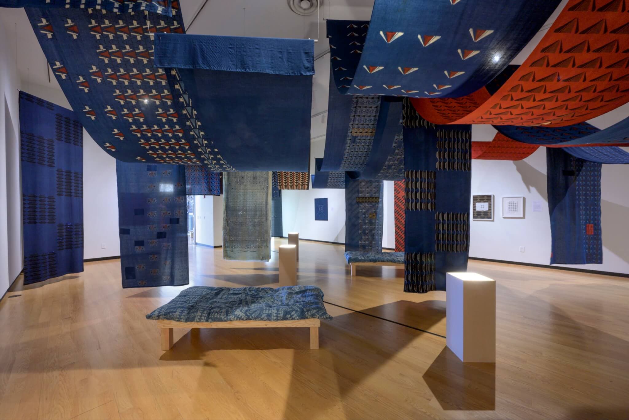 Long bolts of blue and red patterned fabric create a hung structure across a gallery space.